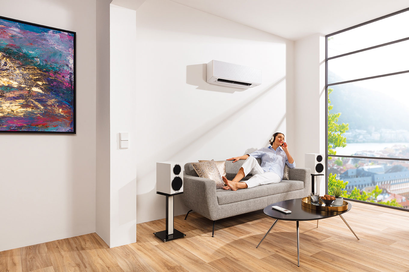 Moderne Wohnzimmerszene mit großer Fensterfront, grauem Sofa, weißer Klimaanlage an der Wand, Frau mit Kopfhörern auf dem Sofa.